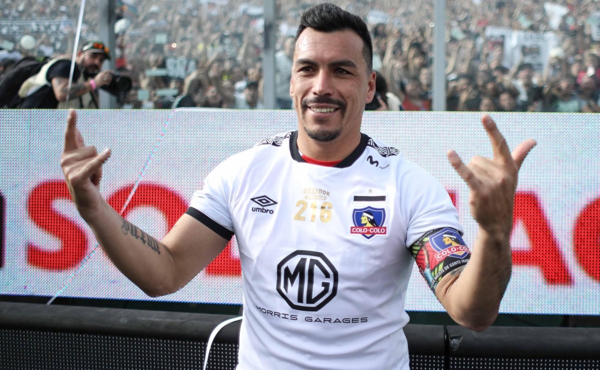 Esteban Paredes posa con la Garra Blanca de fondo y realiza el clásico saludo de Colo-Colo con ambas manos.