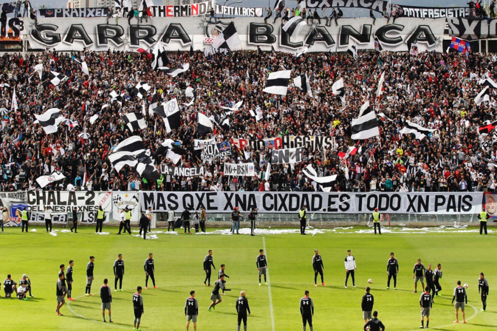 Hinchas de Colo-Colo desplegados en el sector cordillera del Estadio Monumental, observando un entrenamiento de Colo-Colo en la cancha principal del recinto deportivo de Macul. En la parte posterior de la galería se puede observar un lienzo con la consigna "Garra Blanca".