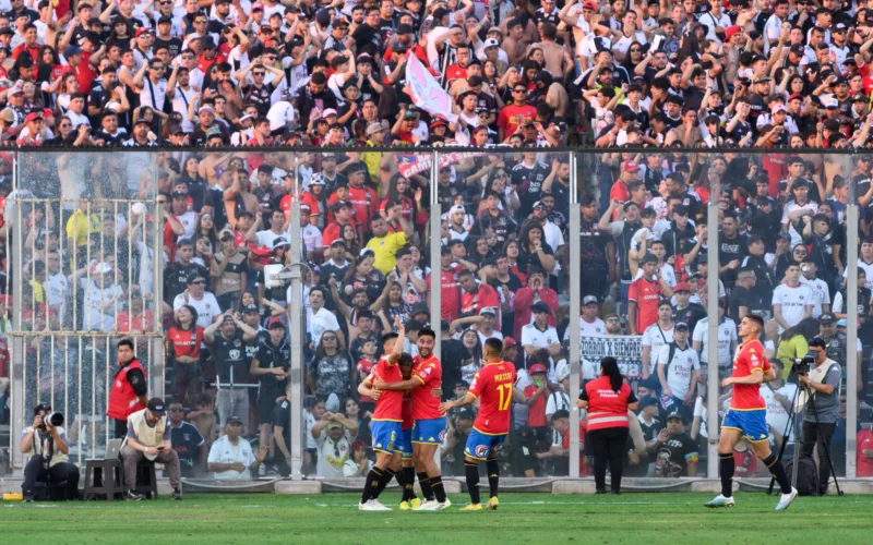 Futbolistas de Unión Española celebran un gol en la cancha principal del Estadio Monumental, mientras que en el fondo aparecen los hinchas de Colo-Colo en el sector Arica.