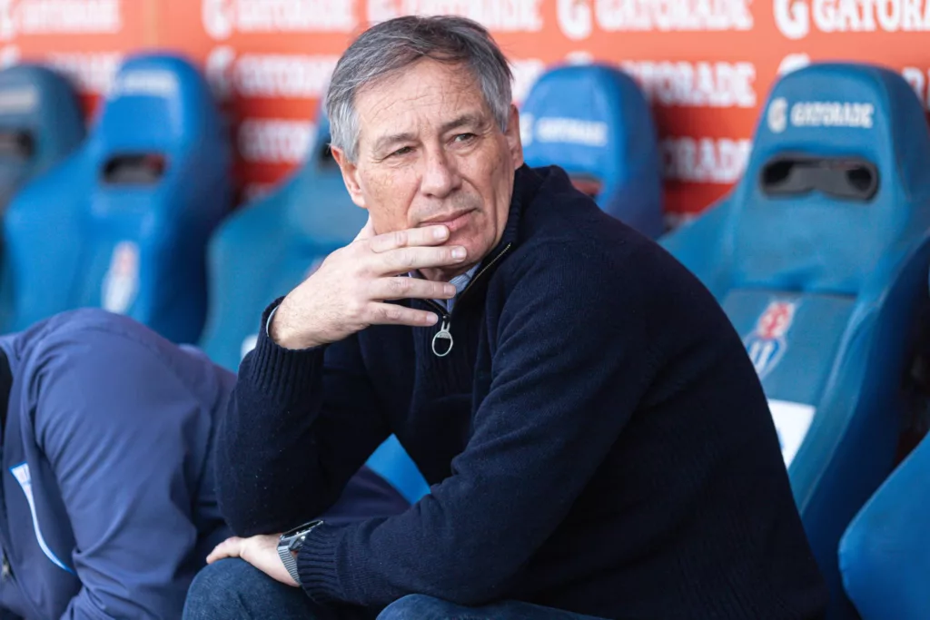 Ariel Holan sentado en un banco de suplentes con el escudo de la Universidad de Chile