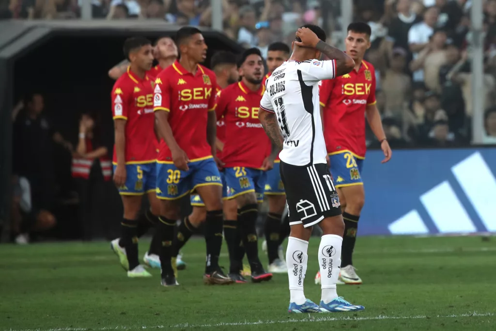 Marcos Bolados tomándose la cabeza mientras jugadores de Unión Española celebran un gol.