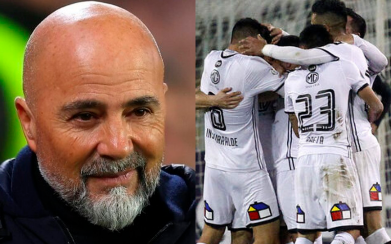 Primer plano al rostro sonriente del entrenador de fútbol profesional, Jorge Sampaoli, mientras que a mano derecha aparecen los jugadores de Colo-Colo celebrando un gol durante la temporada 2018.