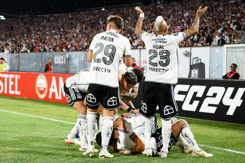 Jugadores de Colo-Colo se abrazan en el suelo y otros levantan sus brazos para alentar al público del Estadio Monumental en pleno partido por Copa Libertadores ante Sportivo Trinidense este 2024.