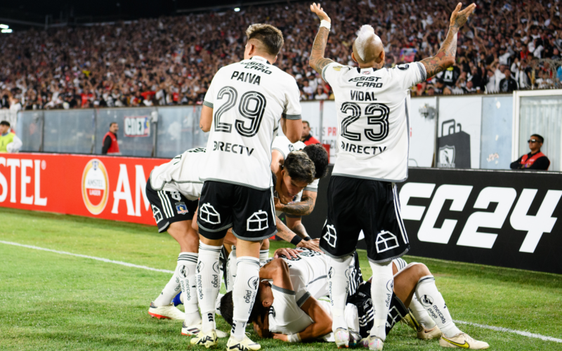 Jugadores de Colo-Colo se abrazan en el suelo y otros levantan sus brazos para alentar al público del Estadio Monumental en pleno partido por Copa Libertadores ante Sportivo Trinidense este 2024.