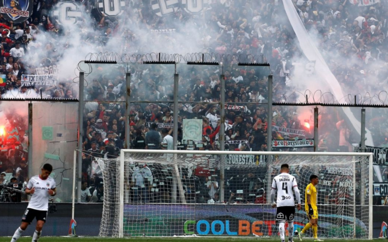 Fotografía a la galería norte del Estadio Monumental, donde se puede ver repleta de hinchas que encienden bengalas y levantan los bombos para alentar a los jugadores de Colo-Colo en pleno partido. En el césped se puede ver en miniatura a Alan Saldivia, Brayan Cortés y Esteban Pavez.