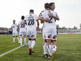 Esteban Paredes abraza a Gabriel Costa tras anotar un gol con la camiseta de Colo-Colo durante la temporada 2019 en el Estadio Municipal de La Cisterna, mientras que el resto de sus compañeros camina al círculo central.