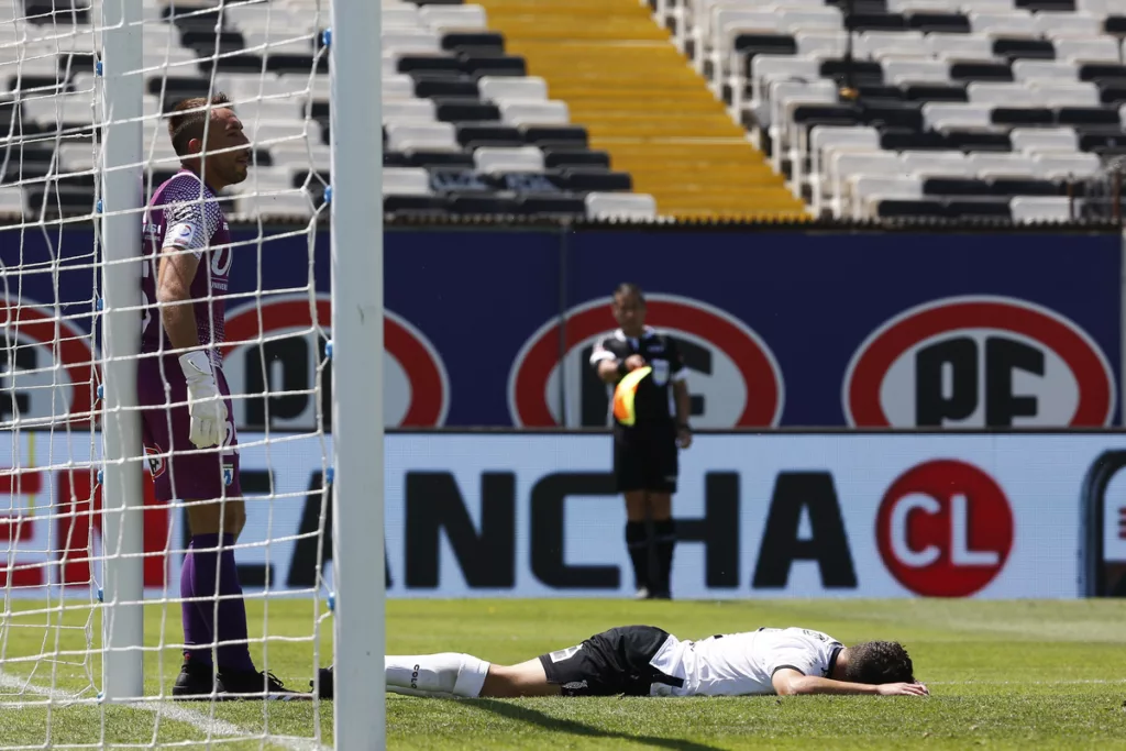 Branco Provoste derramado en el piso en pleno partido con la camiseta de Colo-Colo, mientras que Sebastián Pérez se encuentra parado en el arco norte del Estadio Monumental.