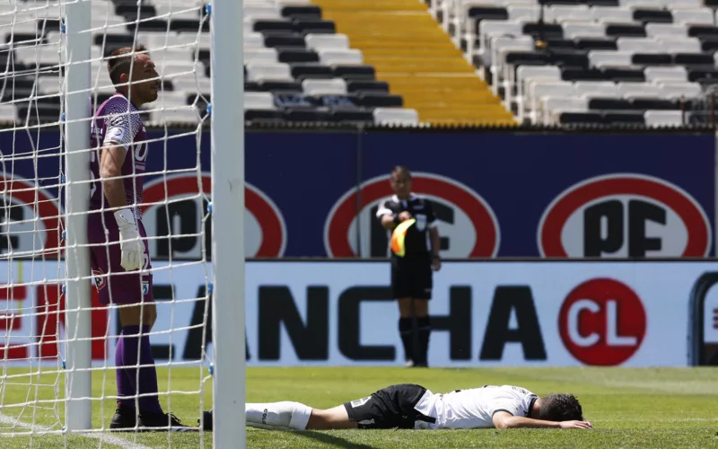 Branco Provoste derramado en el piso en pleno partido con la camiseta de Colo-Colo, mientras que Sebastián Pérez se encuentra parado en el arco norte del Estadio Monumental.