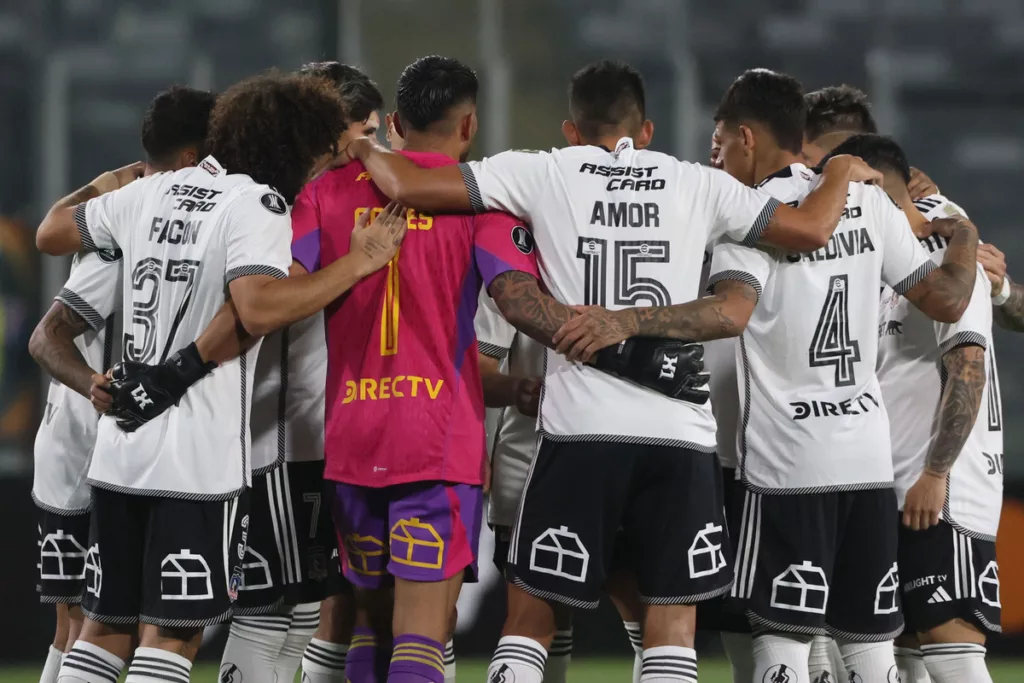 Fotografía a las espaldas de los futbolistas de Colo-Colo masculino durante la temporada 2024, quienes se encuentran abrazados y arengándose antes de jugar un partido.