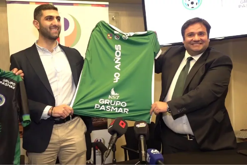 Aziz Mosa junto al presidente de Deportes Puerto Montt levantan una polera del club donde se ve la parte trasera.