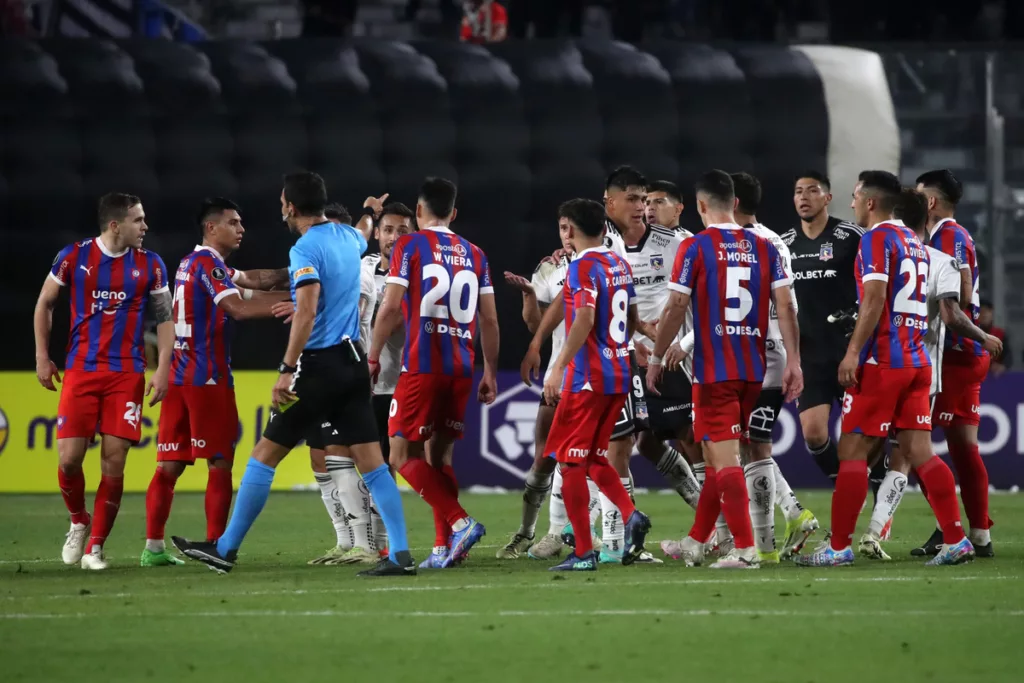 Futbolistas de Cerro Porteño rodean al árbitro Facundo Tello y reclaman un cobro, mientras que los deportistas de Colo-Colo miran atentos a la situación.