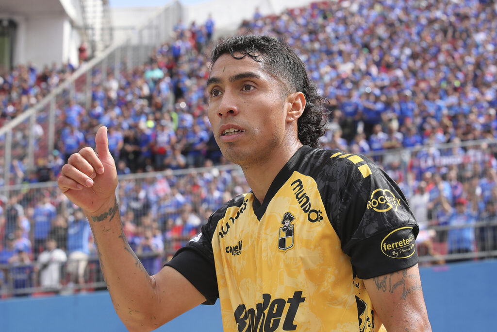 Luciano Cabral levanta su pulgar en pleno partido con l camiseta de Coquimbo Unido y la hinchada de Universidad de Chile de fondo.