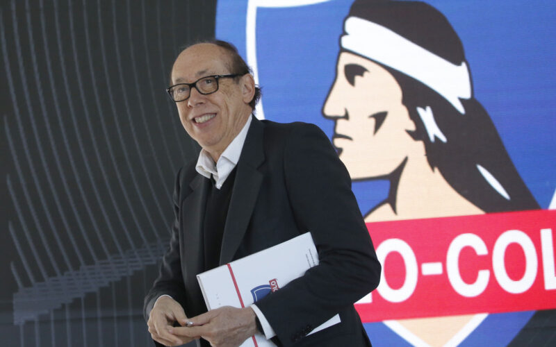 Alfredo Stöhwing sostiene una carpeta en sus manos y de fondo está el logo de Colo-Colo en una pantalla.