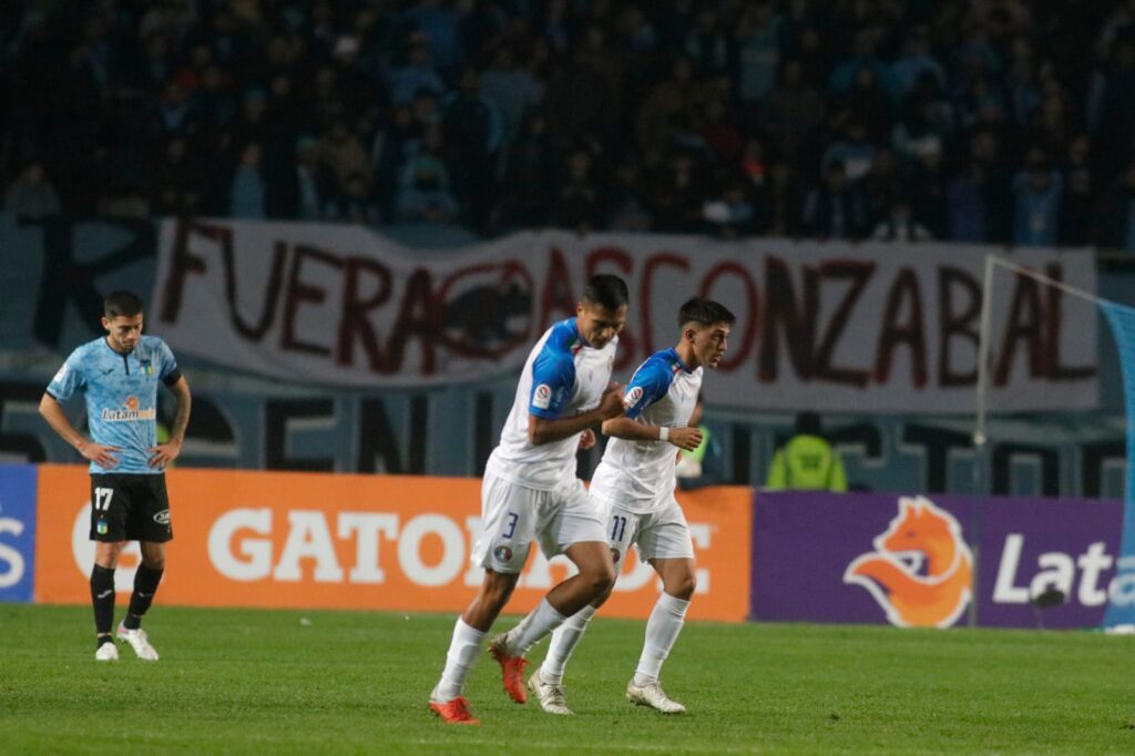 Audax Italiano celebrando un gol con cartel de barra de O'Higgins de fondo.